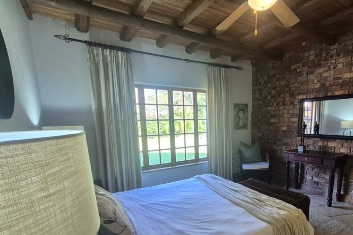 A cozy bedroom featuring an exposed brick feature wall, timber beam ceiling, and tiled flooring with views of the garden.