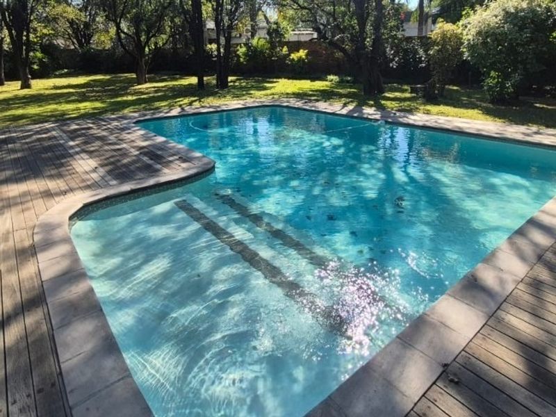 A well-maintained swimming pool area featuring timber decking surround and a lush, mature garden setting.