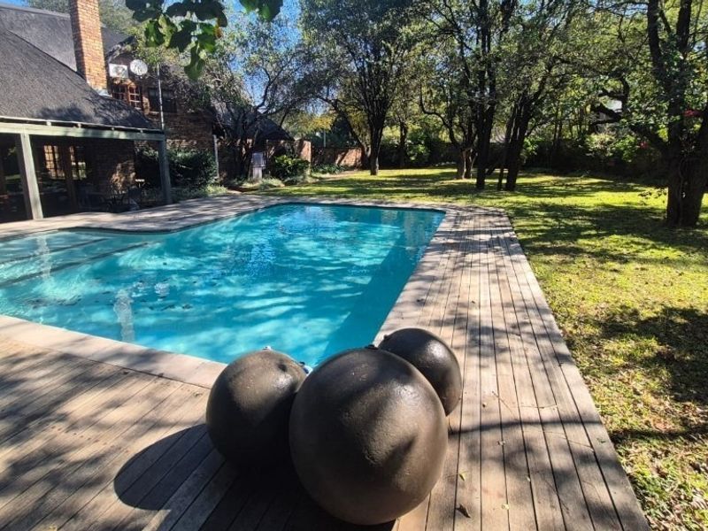 Outdoor pool area featuring a swimming pool, timber decking, and a view of the garden with mature trees and a thatched roof home.