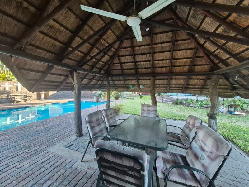 Outdoor entertainment area featuring a thatched lapa with exposed timber beams, ceiling fan, and brick paving, overlooking a swimming pool.