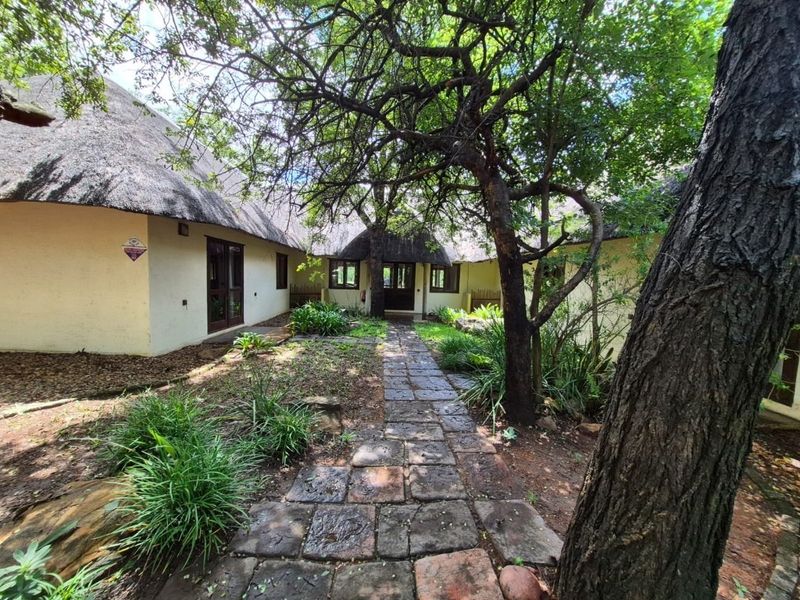 A rear garden view featuring a traditional thatch roof house, stone paved pathway, and mature trees leading to the building entrance.