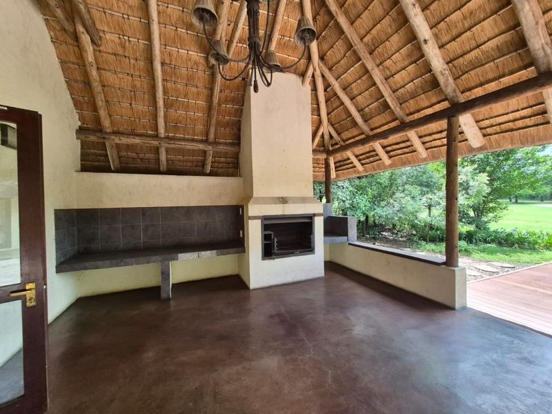 Covered outdoor patio featuring a traditional thatched roof, exposed timber beams, built-in braai, and concrete prep surfaces overlooking the garden.