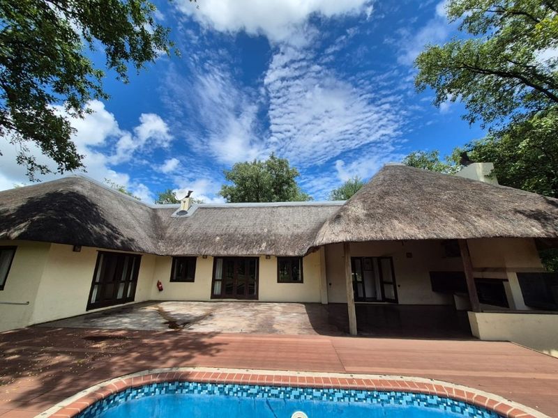 Rear view of a thatched-roof home featuring a swimming pool, timber decking, and a covered patio area with a built-in braai.
