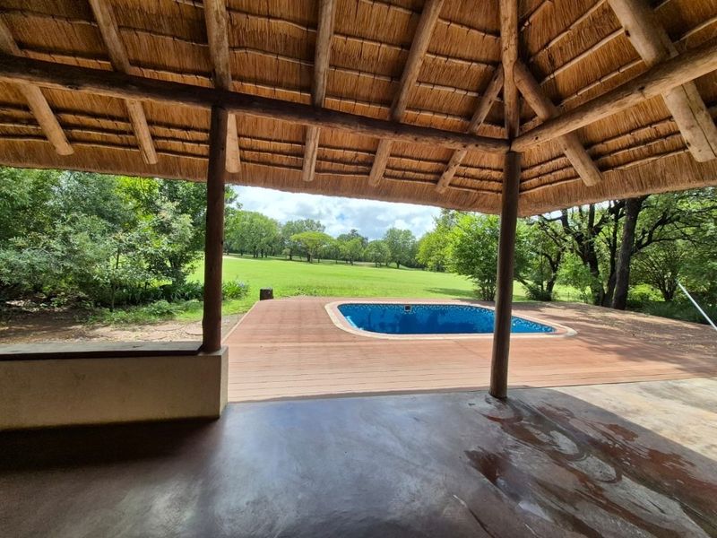 Outdoor entertainment area featuring a thatched roof lapa, wooden decking, and a swimming pool overlooking a lush green garden.