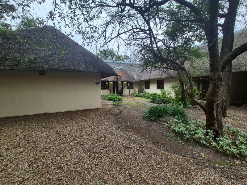 A view of the rear exterior featuring traditional thatch roofing, a gravel courtyard area, and established garden landscaping with mature trees.