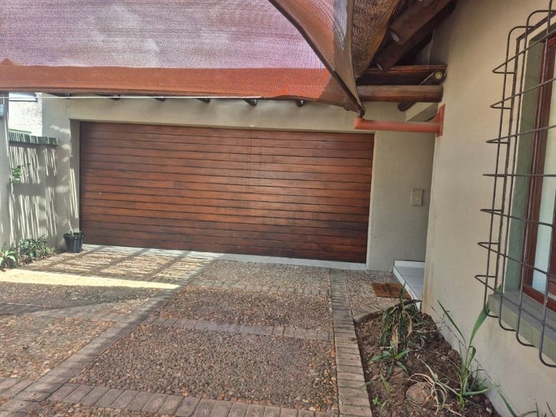 A secure garage area featuring a wooden sectional door, covered by a shade cloth canopy, with an exposed aggregate driveway and security bars.