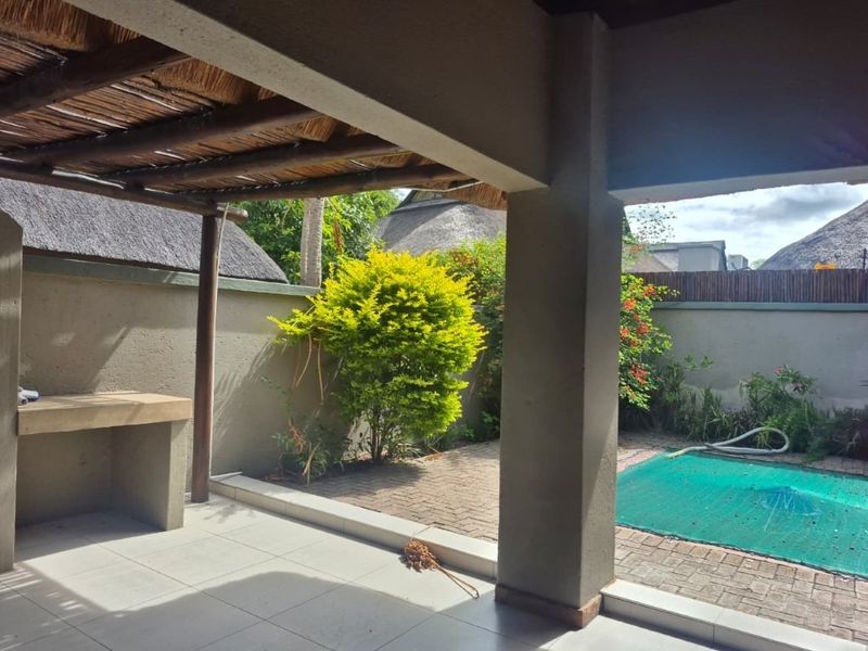 Covered patio area featuring a thatched roof, built-in braai, and tiled flooring, overlooking a paved garden space with a pool.