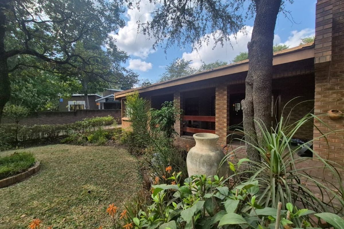 A view of the rear exterior featuring a covered brick patio, established garden, and mature trees.