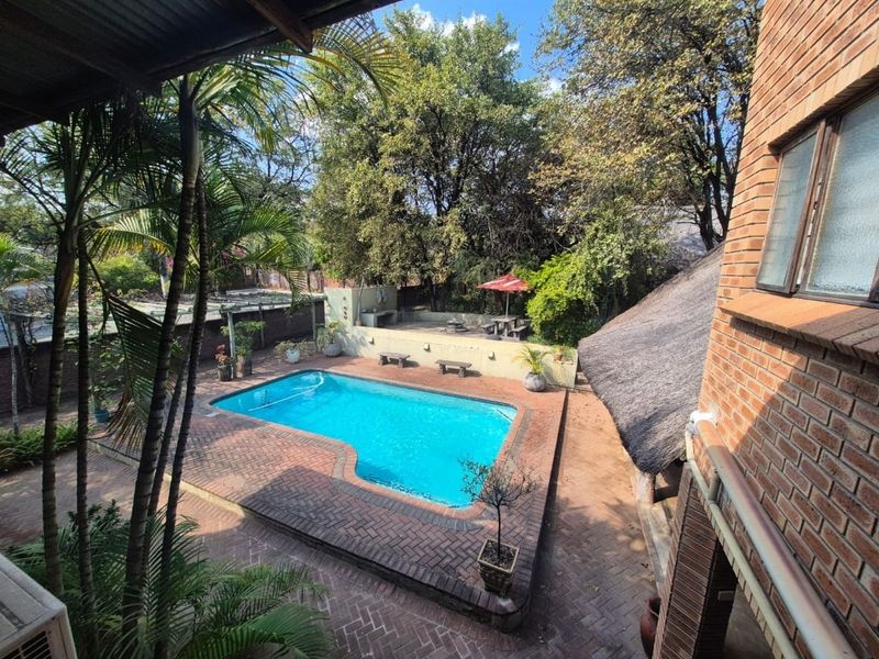 Outdoor entertainment area featuring a swimming pool, brick paving, and a built-in braai, set within a landscaped garden with mature trees.
