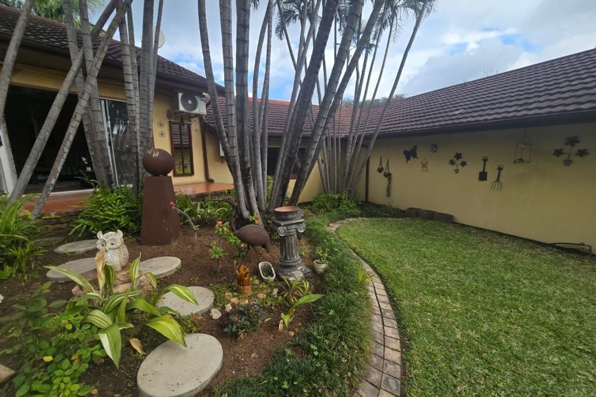 A rear garden area featuring a lawn, landscaped garden beds with stone pavers, and a tiled roof structure with an exterior air conditioning unit.