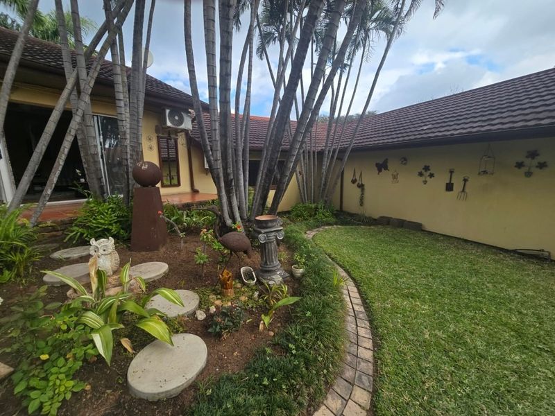 A rear garden area featuring a lawn, landscaped garden beds with stone pavers, and a tiled roof structure with an exterior air conditioning unit.