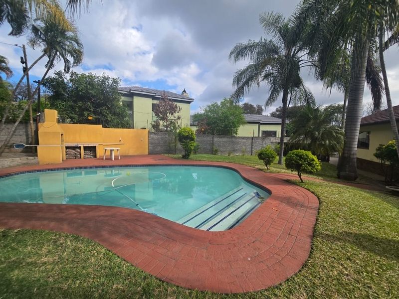 Outdoor pool area featuring a swimming pool, built-in braai, and landscaped garden with mature palm trees and brick paving.