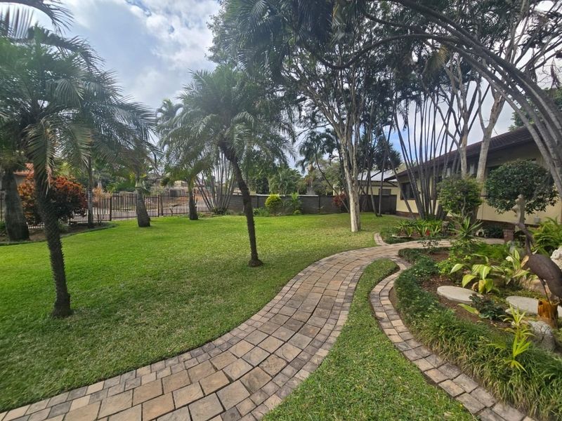 A well-maintained garden featuring a paved walkway, lush green lawn, and mature trees, enclosed by a perimeter wall.