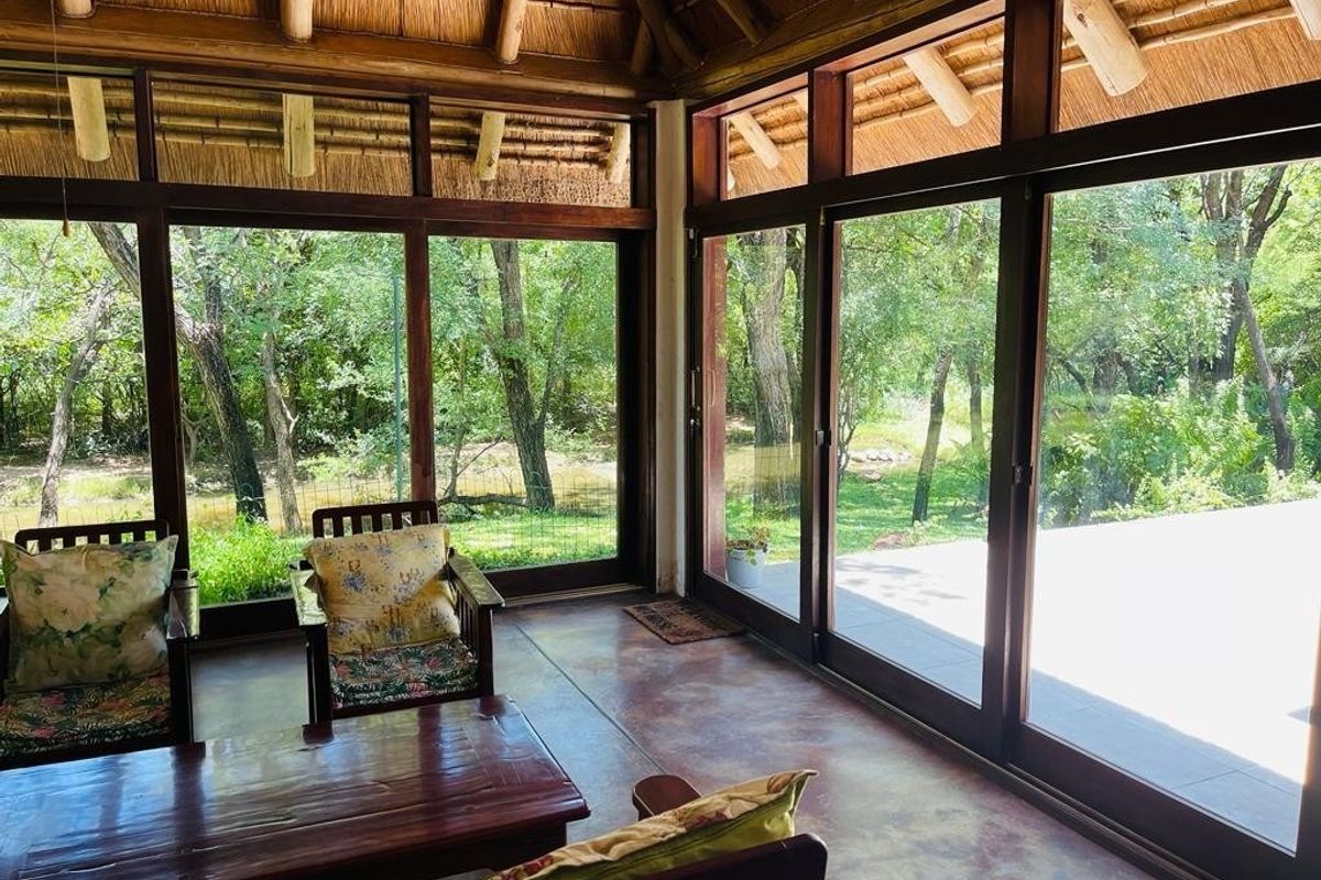 A bright, enclosed patio area featuring a traditional thatched roof, exposed timber rafters, and expansive glass sliding doors overlooking a lush garden setting.