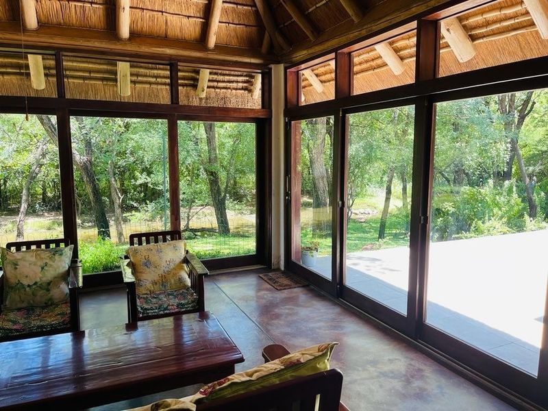 A bright, enclosed patio area featuring a traditional thatched roof, exposed timber rafters, and expansive glass sliding doors overlooking a lush garden setting.
