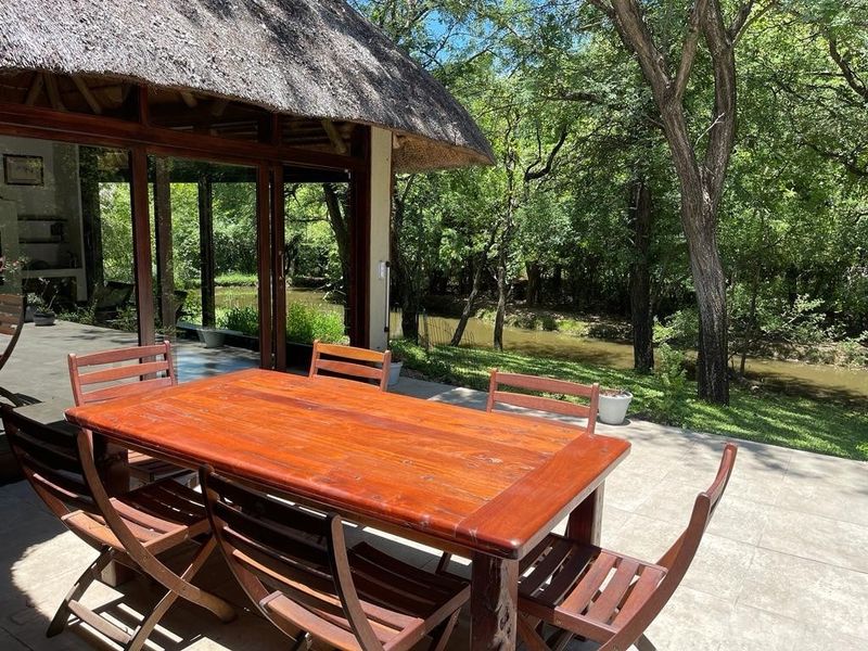 A covered patio area featuring a thatched roof and large sliding glass doors, overlooking a lush garden and river setting.