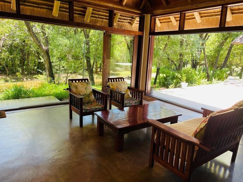 A spacious covered patio featuring a traditional thatched roof, exposed timber beams, and large sliding glass doors that open to a lush garden view.