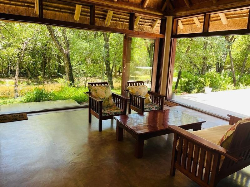 A covered patio area featuring a thatched roof, exposed timber beams, and large sliding glass doors that open to a lush garden view.