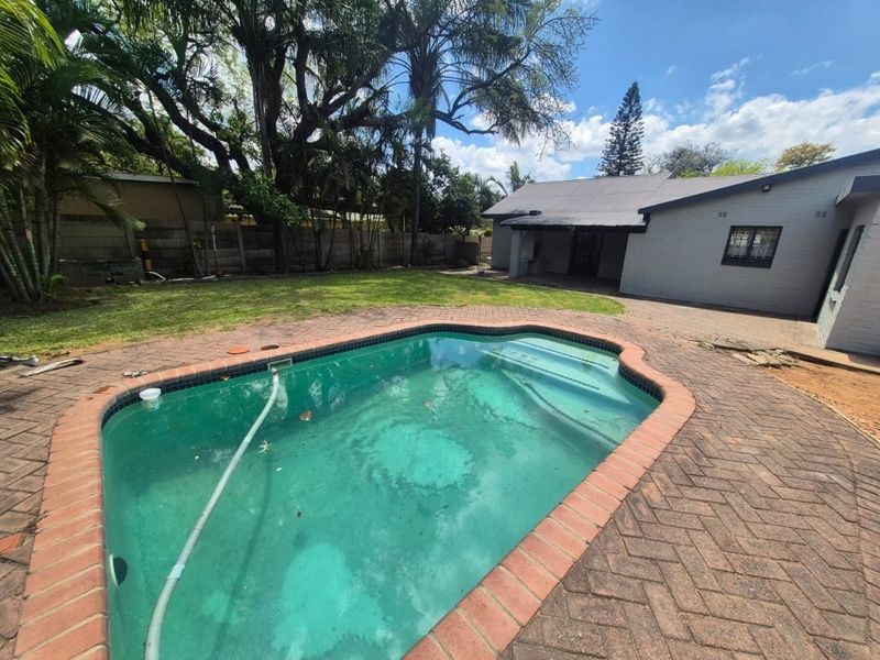 Outdoor pool area featuring a swimming pool, brick paving, and a covered patio, set within a garden with mature trees and boundary walls.
