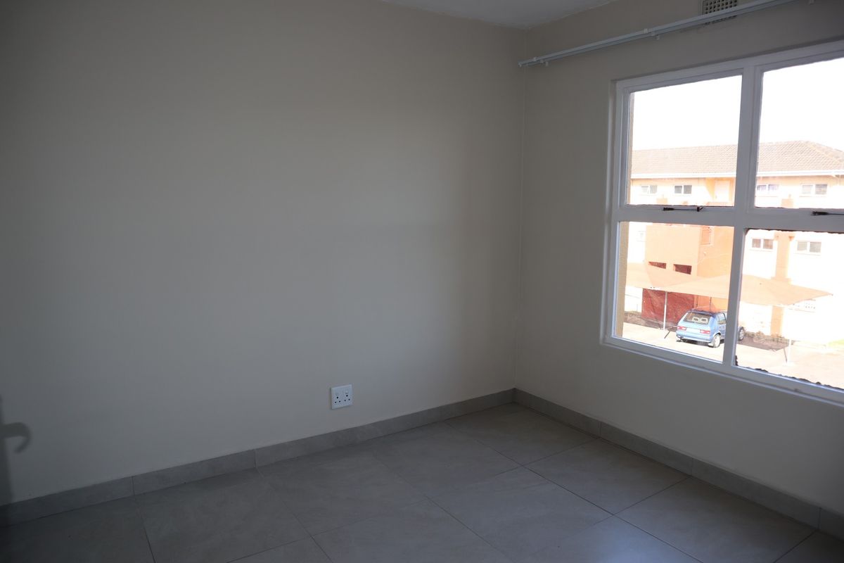 A bright, empty bedroom featuring neutral wall paint, tiled flooring, and a large window providing natural light and a view of the complex exterior.