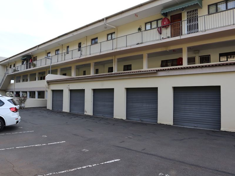 Exterior view of an apartment complex featuring a row of roller shutter garages, an asphalt parking area, and upper-level balconies with metal railings.