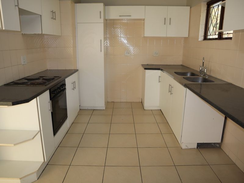 A functional, empty kitchen featuring white cabinetry, dark laminate countertops, a built-in electric oven and hob, and neutral tiled splashbacks and flooring.