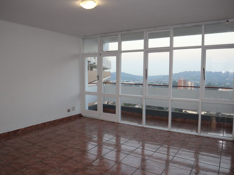An empty living room featuring tiled flooring and a large glass window wall with access to a balcony, offering elevated urban views.
