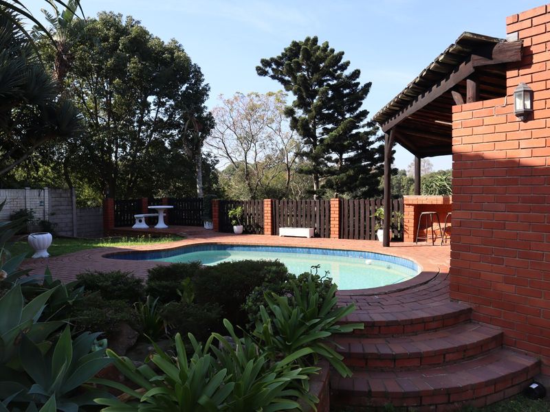 Outdoor pool area featuring a brick-paved patio, a covered entertainment space with a built-in brick bar, and established garden surroundings.