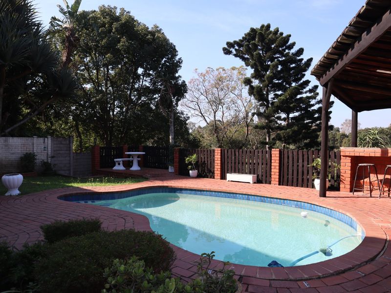 Outdoor pool area featuring brick paving, a covered patio with a built-in brick braai, and established garden surroundings.