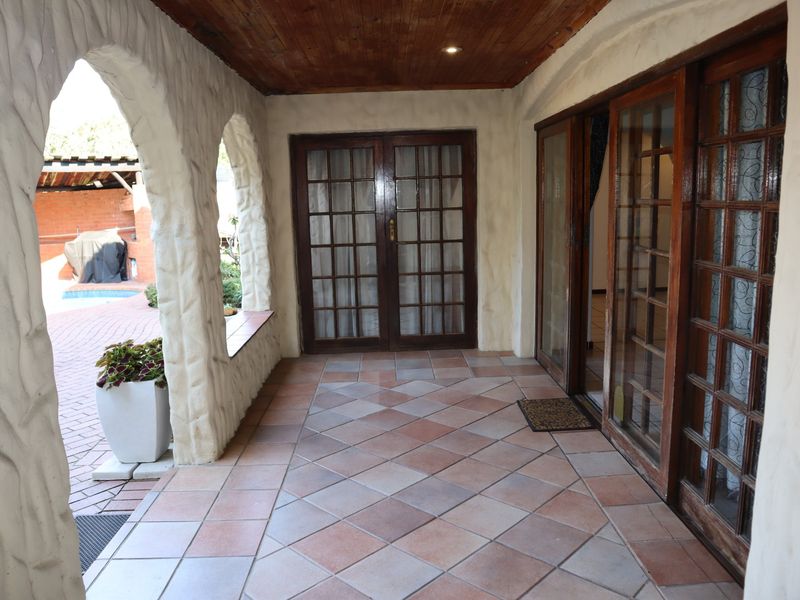 A covered patio area featuring tiled flooring, a timber ceiling, and arched masonry pillars, providing access to the interior through timber-framed glass doors.