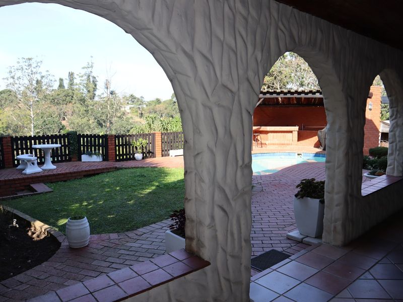 Covered patio featuring distinctive arched masonry columns, overlooking a landscaped garden with brick pathways, a swimming pool, and a built-in outdoor bar area.