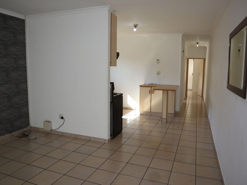 Open-plan living area featuring tiled flooring, a compact kitchen corner with built-in cabinetry, and a breakfast counter, leading into a hallway.
