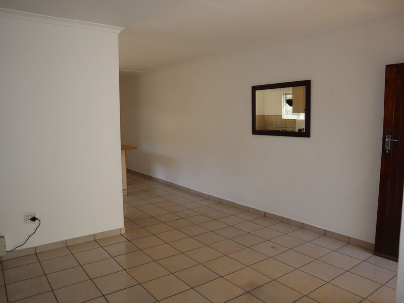 An empty living area featuring neutral tiled flooring, a wooden door, and a view through to the kitchen area.