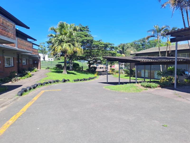 A view of the rear exterior area featuring a paved driveway, covered carport parking, and established garden landscaping between residential flat blocks.