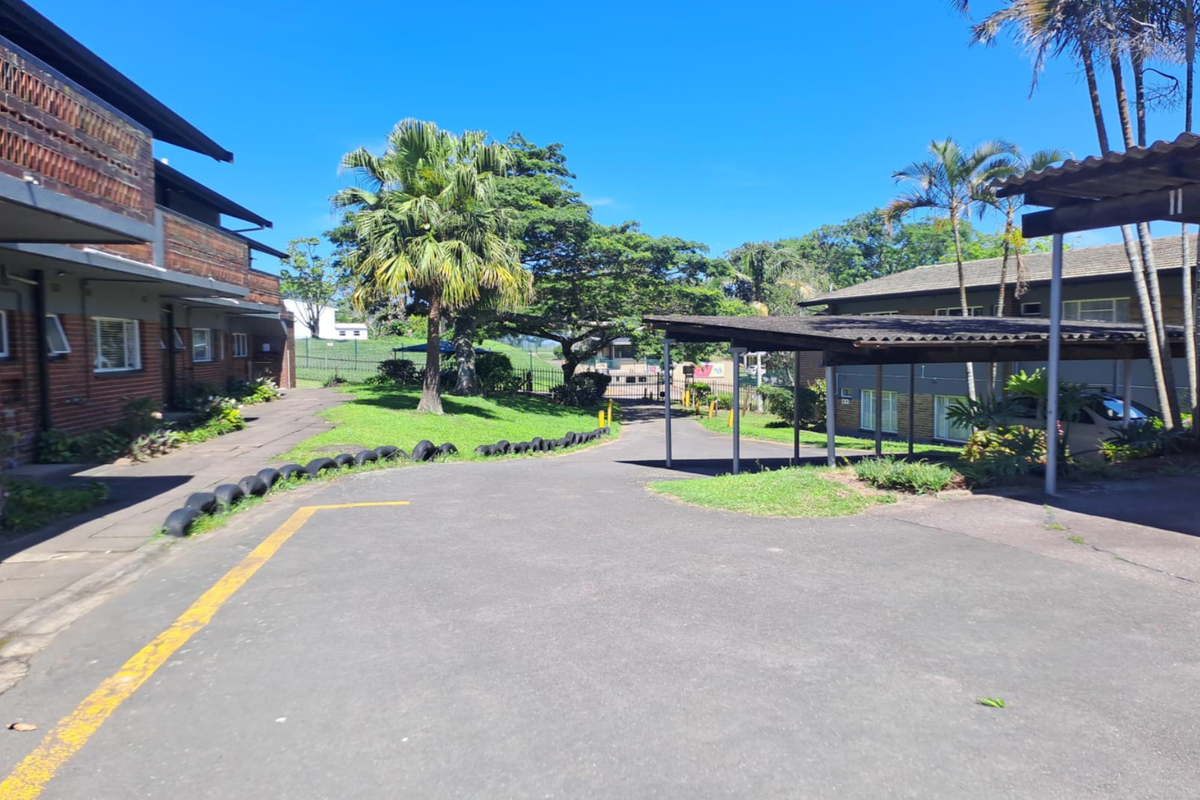 A view of the rear exterior area featuring a paved driveway, covered carport parking, and established garden landscaping between residential flat blocks.