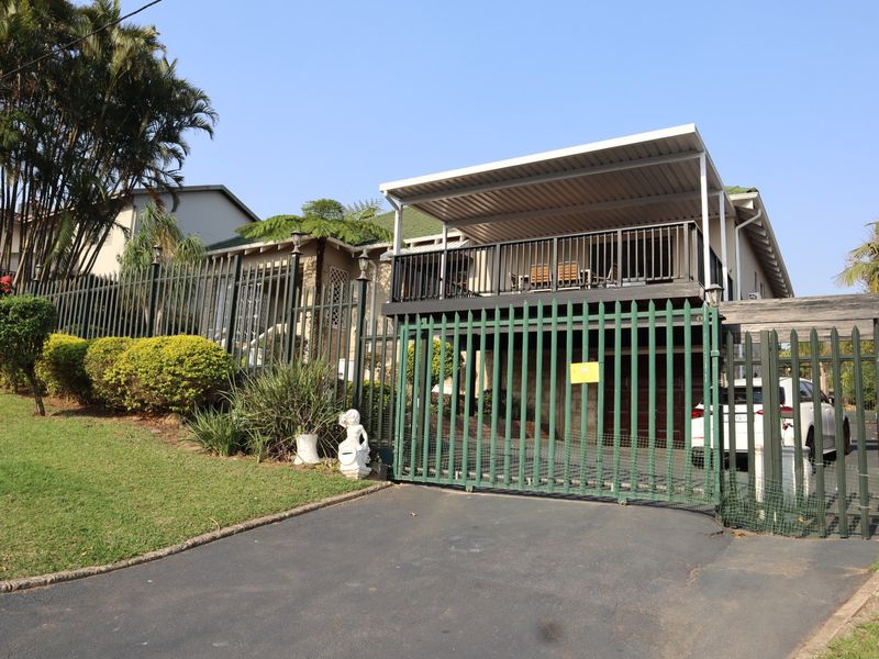 Front view of the property featuring a secure gated entrance, asphalt driveway, and an elevated covered balcony with metal railings.