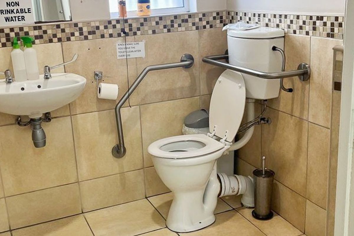 Accessible commercial bathroom featuring a wall-mounted basin, toilet, and safety grab rails with neutral tiled finishes.