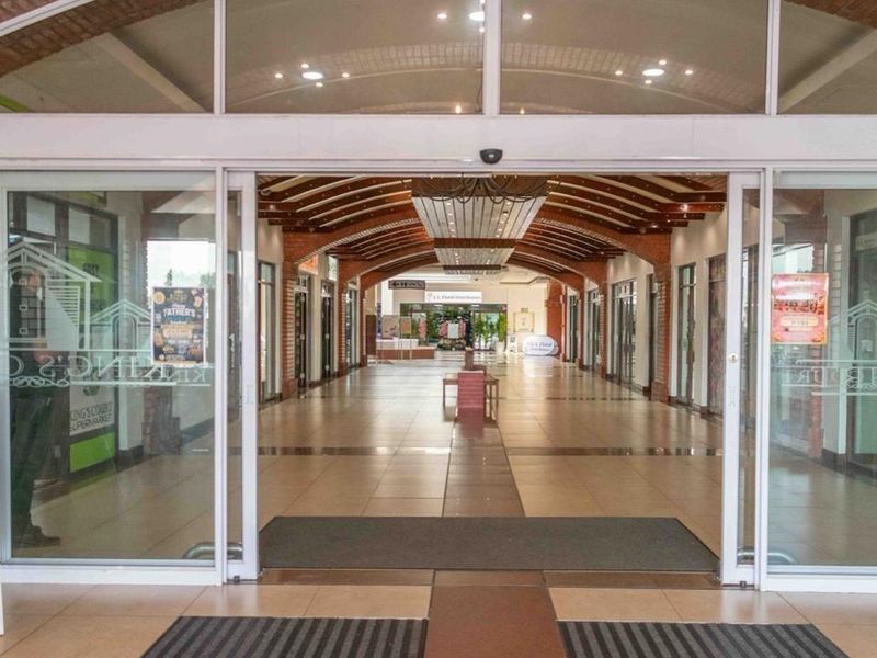 A bright, welcoming commercial entrance featuring automatic sliding glass doors, decorative brick archways, and a spacious tiled walkway leading into the shopping center.
