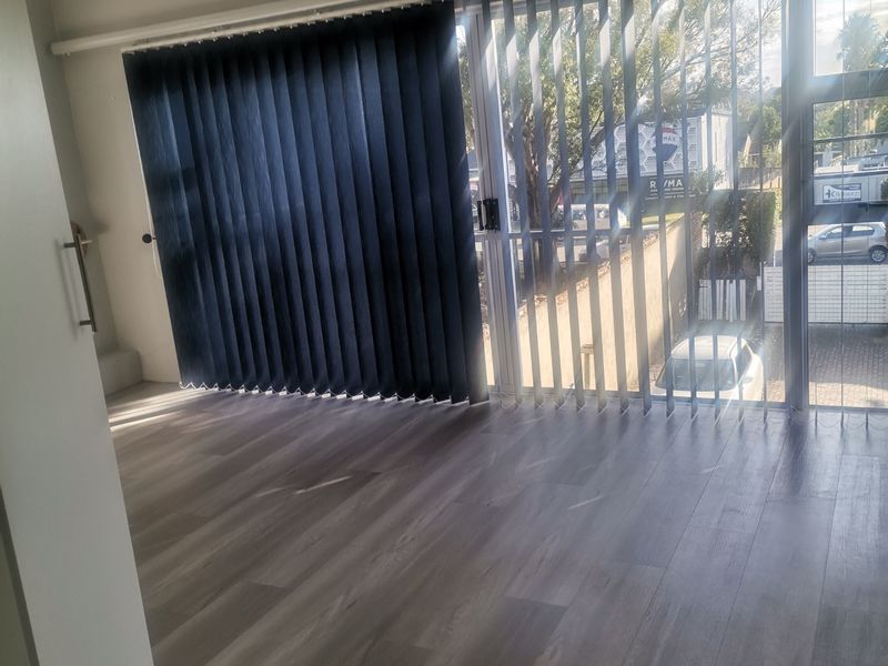 A bright, empty office space featuring modern laminate flooring, large glass sliding doors with vertical blinds, and a ceiling-mounted light fixture.
