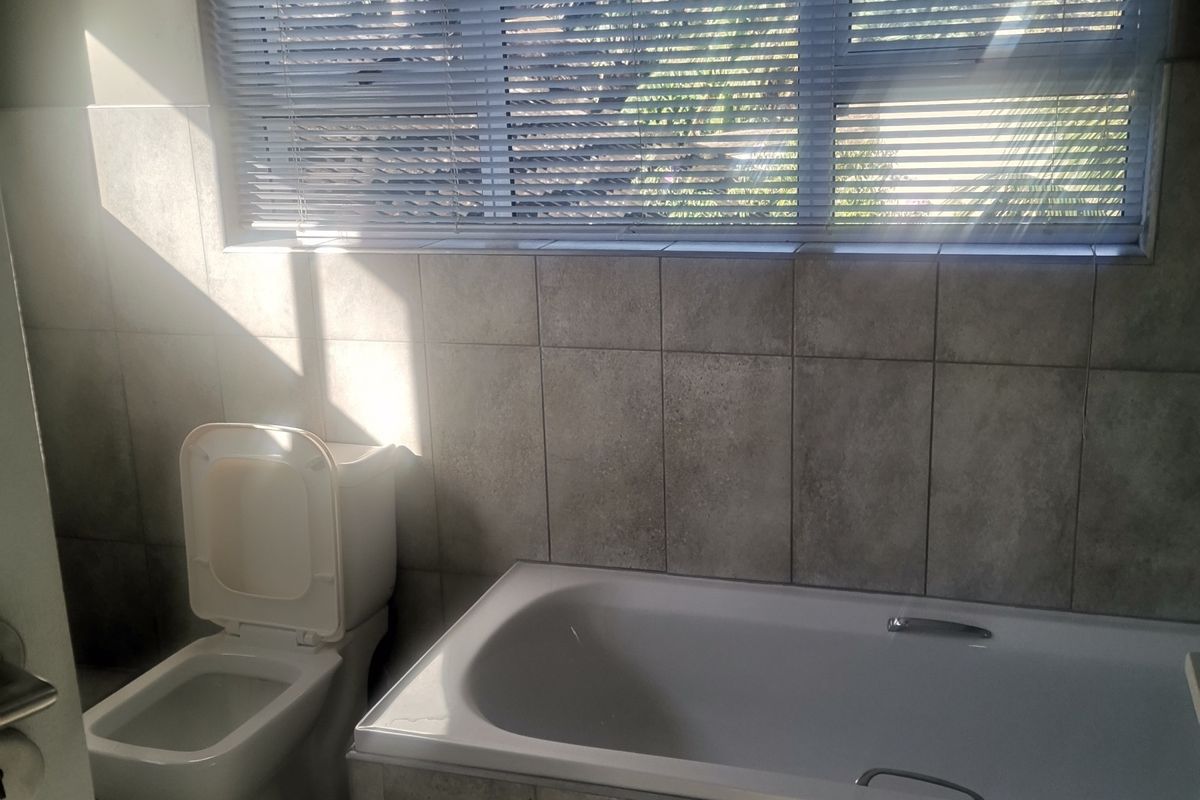 A clean, functional bathroom featuring a built-in bathtub, toilet, and neutral wall and floor tiling with natural light from a large window.