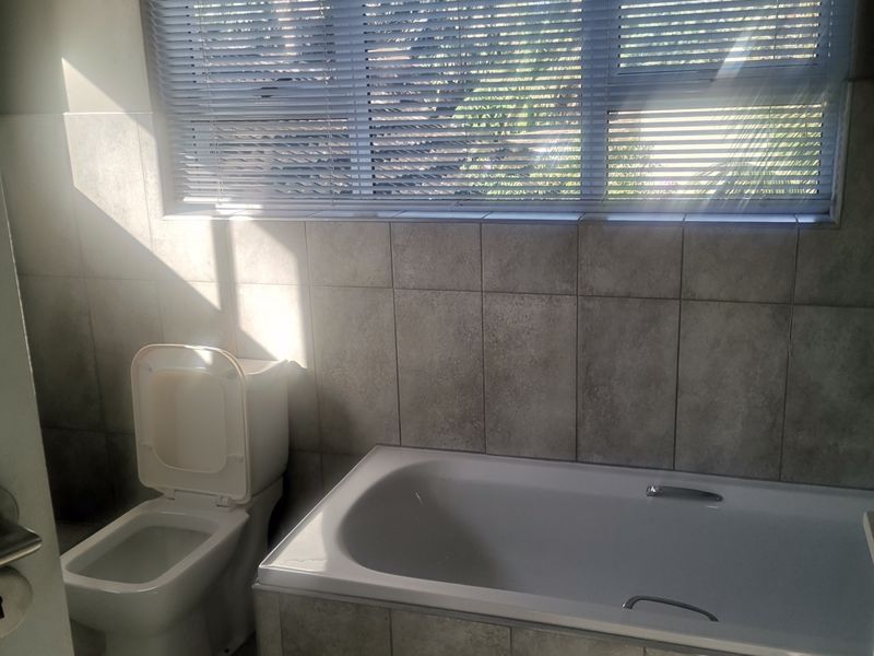 A clean, functional bathroom featuring a built-in bathtub, toilet, and neutral wall and floor tiling with natural light from a large window.