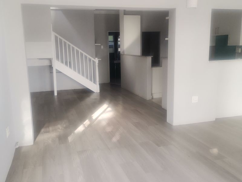 An open-plan living area featuring light-toned laminate flooring, a staircase with a white balustrade, and a pass-through window connecting to the kitchen area.
