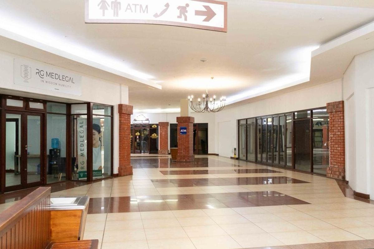 A spacious, well-lit retail hallway featuring tiled flooring, decorative brick columns, and glass-fronted commercial units with clear directional signage.
