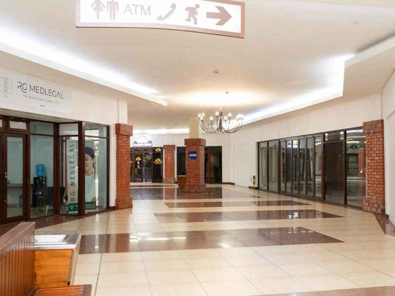 A spacious, well-lit retail hallway featuring tiled flooring, decorative brick columns, and glass-fronted commercial units with clear directional signage.