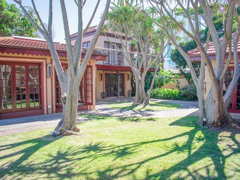 A well-maintained rear garden area featuring mature trees, paved pathways, wooden decking, and multiple access points to the guest house through wooden framed glass doors.