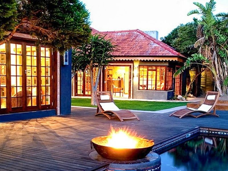 A beautifully landscaped pool area featuring extensive wooden decking, a fire pit, and direct access to the guest house through timber-framed glass doors.