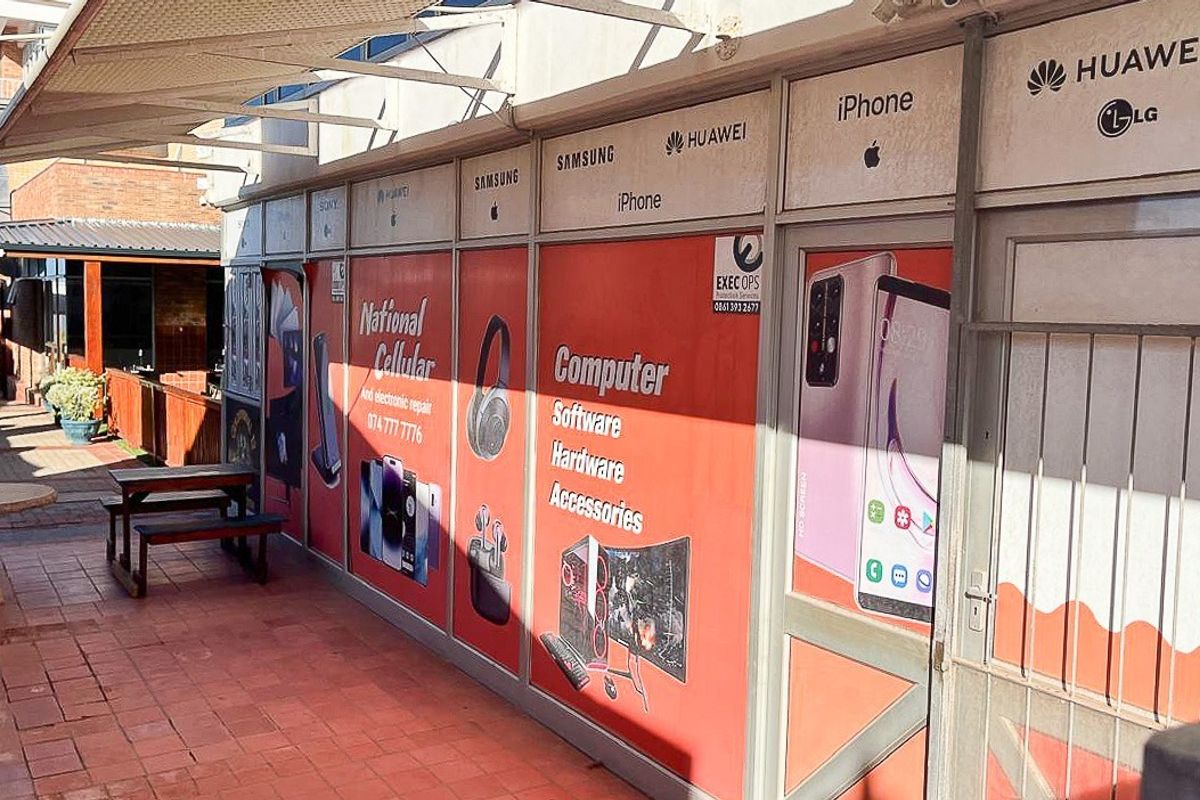 Retail storefront featuring large glass display windows with branding, metal security gate, and a paved walkway area under a covered awning.