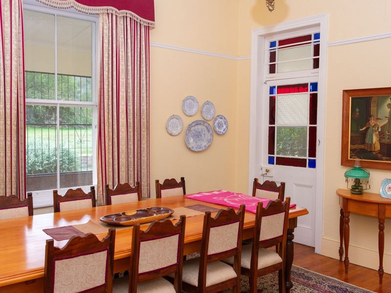 A formal dining room featuring classic timber flooring, a sash window with decorative curtains, and a characterful door with stained glass inserts.