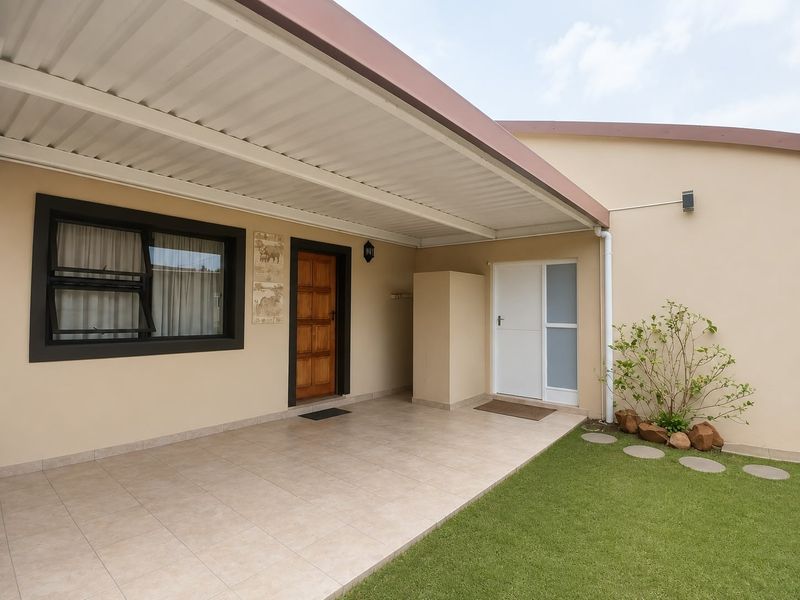 A well-maintained covered patio area featuring tiled flooring, a timber entrance door, and an adjacent small garden space.