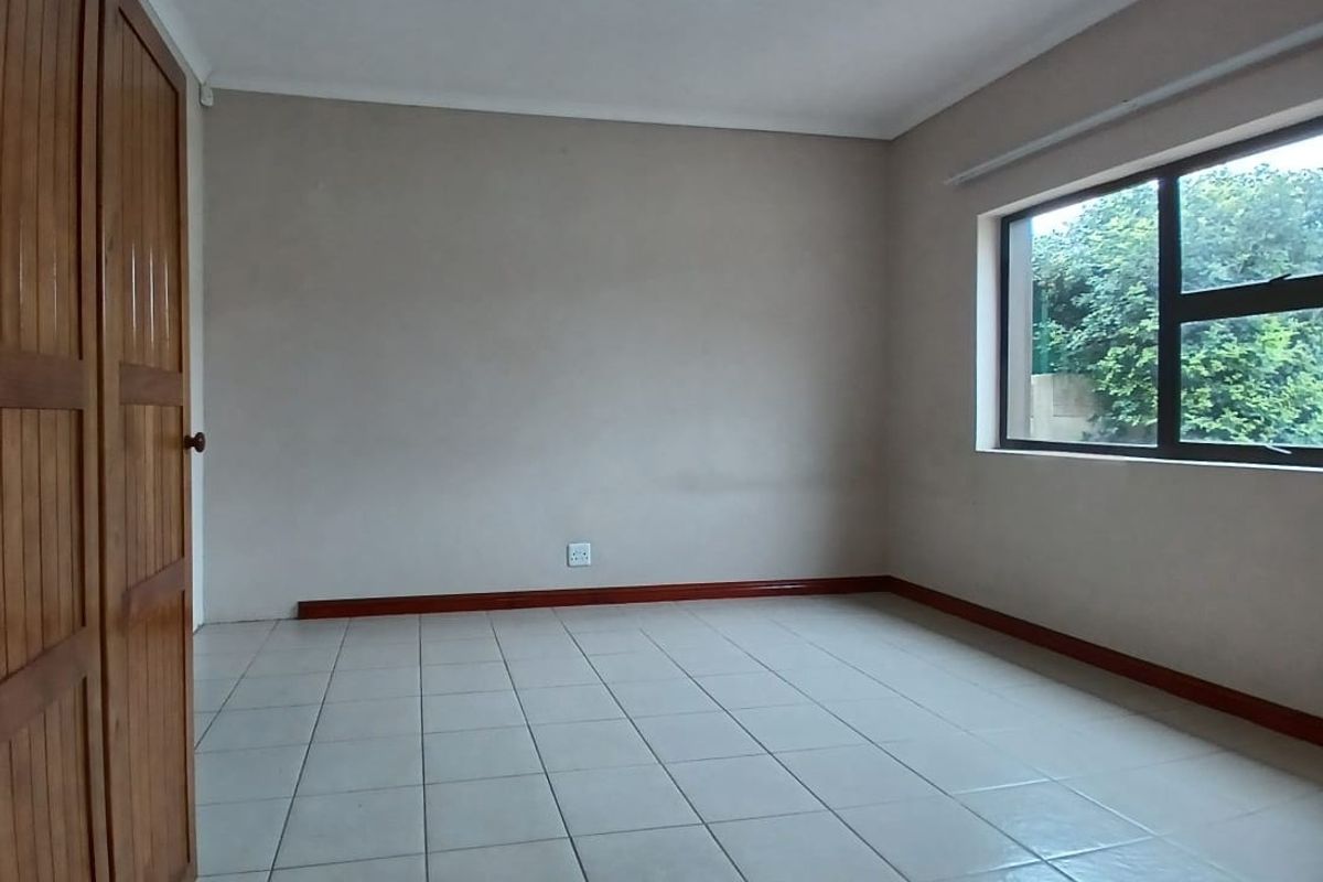 A clean, empty bedroom featuring neutral wall paint, ceramic floor tiles, and built-in wooden cupboards with natural light from the window.